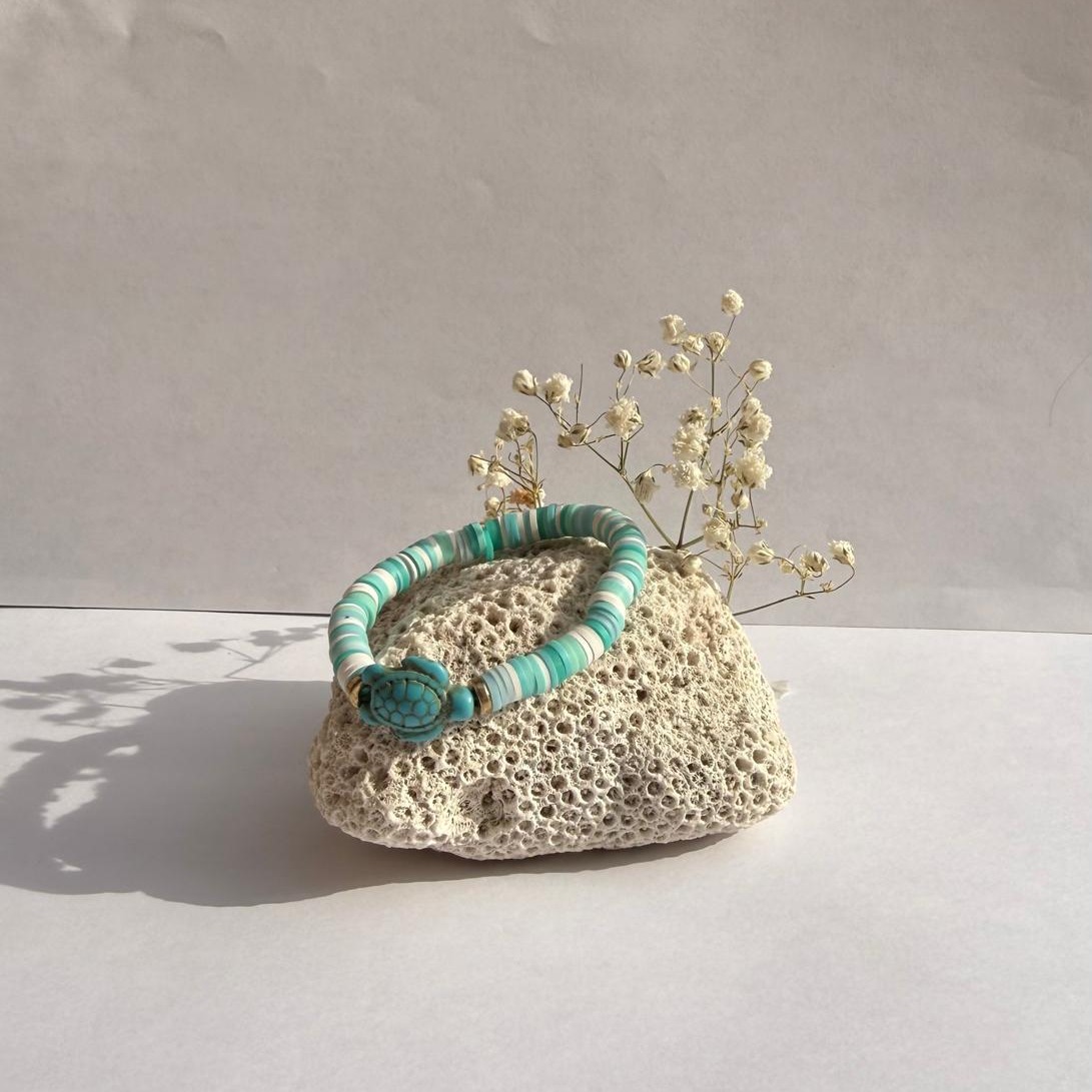 Teal Pop bracelet with a teal turtle charm on a rock with flowers against a white wall