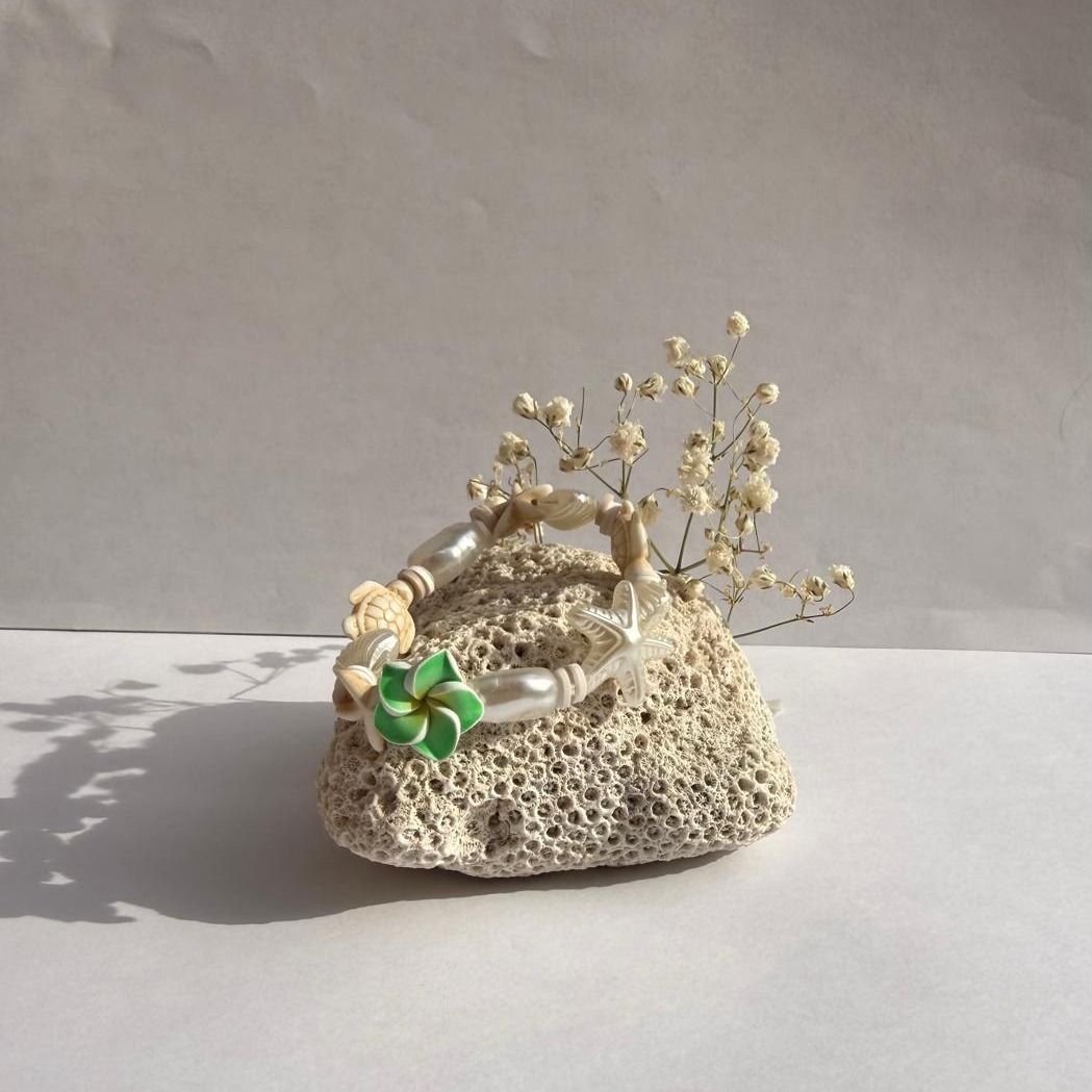 Turtle Blossom bracelet with a green flower charm on a rock with flowers against a white wall